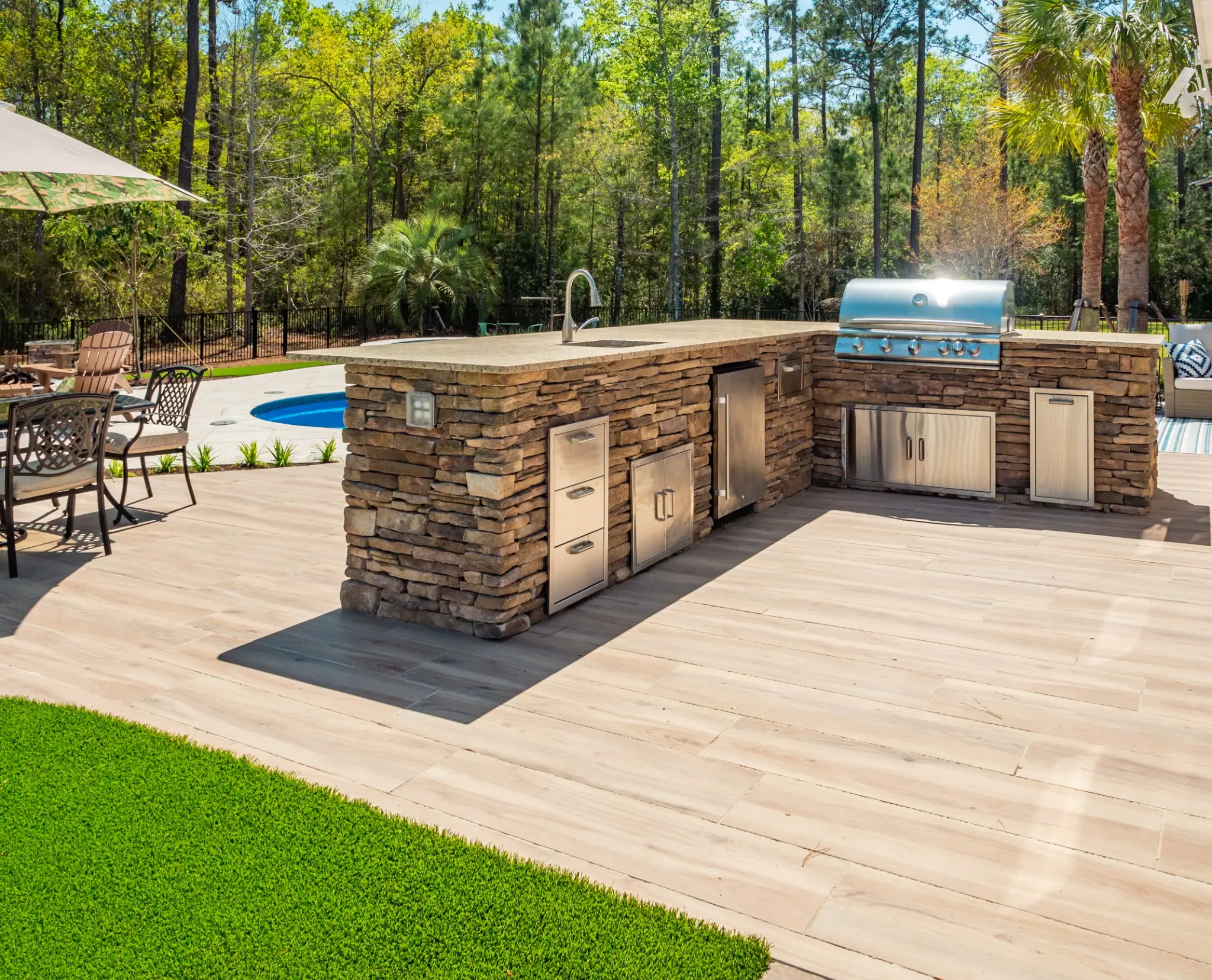 Stone outdoor kitchen with built-in grill, sink, refrigerator, drawers, and poolside countertop
